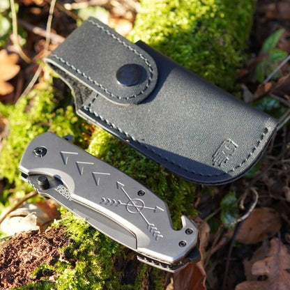 Opgevouwen zakmes met pijlmotief naast zwart leren foedraal op mosrijke ondergrond in natuur