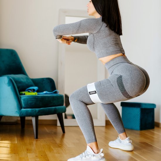 Vrouw in grijze sportkleding doet squats met weerstandsband in lichte woonkamer