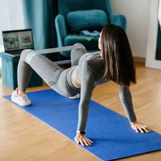 Vrouw doet heupbrug oefening met weerstandsband op een blauwe yogamat in huiselijke omgeving