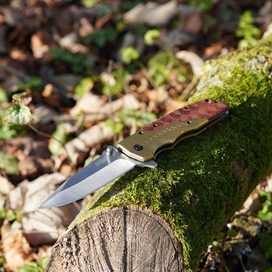 Vouwmes met houten handgreep en stalen lemmet liggend op met mos bedekt boomstam in bosrijke omgeving