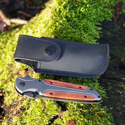 Opgevouwen zakmes met metalen en houten handgreep naast een zwarte leren hoes op mosgrond.