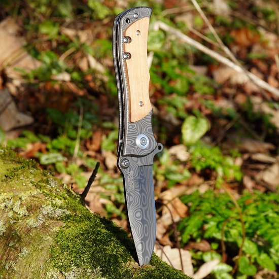 Opvouwbaar mes met houten handgreep en decoratief zwart lemmet geplaatst in mosrijke omgeving.