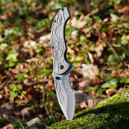 Gesloten mes met grijzig handvat en gedetailleerd reliëf in een bosrijke omgeving.