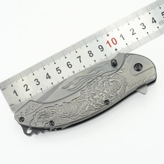 gesloten metalen zakmes met gedetailleerd draakpatroon en liniaal op witte achtergrond