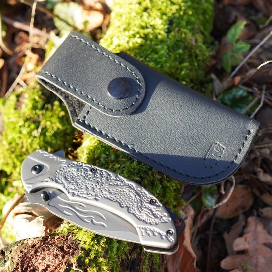 Grijs zakmes met reliëf in krokodillenpatroon naast zwarte leren hoes op mosrijke ondergrond in bos.